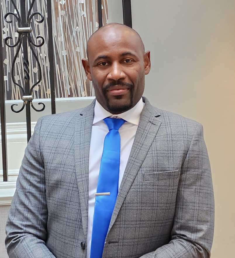 A medium shot Portrait of Sheldon Edwards wearing a grey suit, white shirt and a blue tie